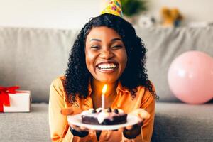 Happy Birthday To You. Cheerful Black Lady In Festive Hat Holding B-Day Cake With Burning Candle Smiling To Camera Sitting At Home. Holiday Celebration, Joyful Events Concept photo