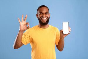 I recommend this new mobile app. Cool young African American guy showing empty smartphone and gesturing okay on blue studio background, mockup for design. Space for advertising website on screen photo