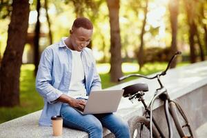 Distance work concept. Joyful African American man with bicycle working on laptop computer at urban park, empty space. Millennial black freelancer doing remote project outside photo
