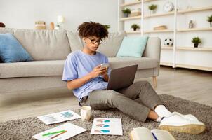 Full length of focused black teen guy using smartphone and laptop for online learning at home. African American teenager chatting with his fellow students or tutor on web, taking part in remote lesson photo