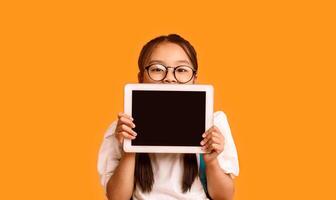 Online Class. Japanese Schoolgirl Showing Digital Tablet Blank Screen Recommending Educational Application Standing Over Yellow Studio Background photo