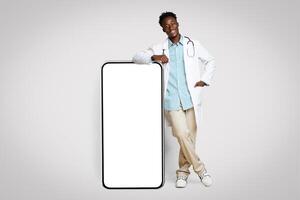 Medical App. African American Male Doctor In Uniform Leaning At Big Blank Smartphone With White Screen, Demonstrating Free Copy Space For Advertisement, Standing On Beige Background, Mockup photo