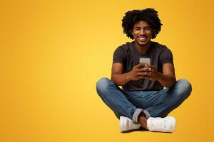 Happy millennial african american guy using smartphone while sitting on floor over beige background in studio, cheerful black male messaging on mobile phone or browsing internet, copy space photo