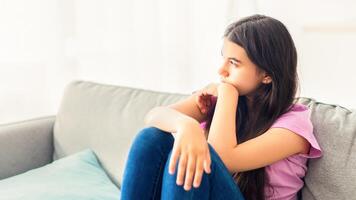 Stressed Indian teenage girl sitting on sofa at home, looking aside, feeling upset and lonely, suffering from lack of communication during covid lockdown. Teen depression concept photo