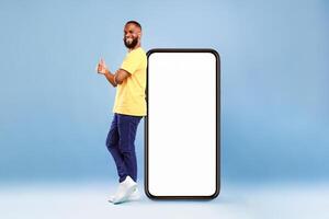 Funky African American guy leaning on giant cellphone, showing thumb up gesture, recommending new mobile app or cool website, mockup for your ad design. Blue studio background photo