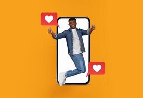 A person joyfully jumps, making a thumbs-up gesture in front of a bright orange background. Large speech bubbles with heart symbols depict excitement in digital communication on a smartphone. photo