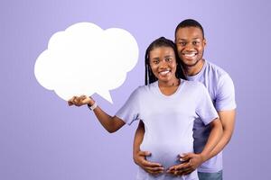 Portrait of smiling black pregnant woman holding empty speech bubble, guy hugging her from behind, couple standing over purple violet studio background, thinking. Free place for text, copy space photo