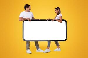 Angry young couple compete fighting over big smartphone, taking away blank white cell screen on orange background, mockup for new mobile app, advertisement or website, withdraw. Online sale template photo