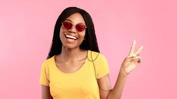 Joyful Black Woman Gesturing Victory Sign To Camera Smiling Posing Over Blue Background, Wearing Sunglasses. Studio Shot Of Happy African American Female Showing Peace Or V-Sign Concept photo