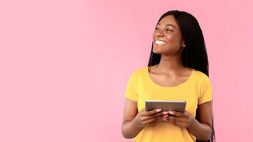 Happy black woman using digital tablet computer looking aside standing over blue studio background. Female browsing internet, looks at copy space. People and gadgets, e-learning. Panorama photo
