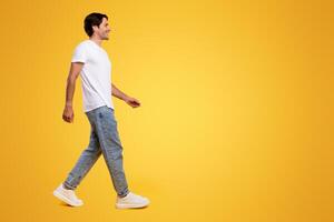 Full length body size profile side view of happy excited bearded guy in casual white t-shirt and jeans walking looking aside at free empty space isolated over orange color background, panoramic banner photo