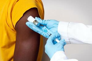 Unrecognizable Doctor Wearing Blue Gloves Making Vaccination Shot To Black Male Patient In Hospital, Medical Worker Using Syringe, Taking Dose Of Vaccine From Vial, Cropped Image, Closeup Shot photo