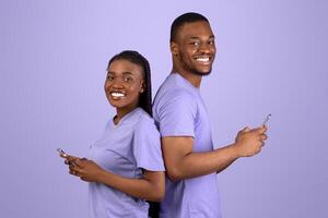 Modern Communication. Profile side view portrait of cheerful positive black couple standing back to back, holding and using their mobile phones and chatting online, looking at camera, violet wall photo