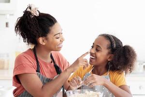 Cheerful black mother and kid in aprons having fun while baking, cute african american mom and daughter playing with flour while making pastry dough at kitchen, enjoying time together, copy space photo
