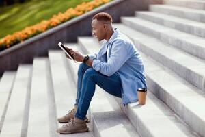 Side view of young black man with takeout coffee using tablet on stairs outdoors. Millennial African American guy with touch pad browsing web, playing online game, sharing post on social media photo