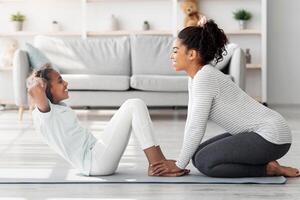 Black mother and daughter in sportswear exercising together at home, sitting on fitness mat and working on abdominal muscles, doing crunches, side view. Sporty family mom and kid doing work out photo