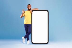 Positive African American guy showing okay gesture next to huge smartphone on blue studio background, empty space for mobile app design on screen. Website or advertisement mockup photo
