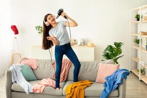Full length of Indian teen girl wearing headphones, using blowdryer as microphone, singing song and dancing on sofa in messy room. Having fun and domestic entertainments during covid lockdown photo