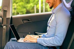 Young arab man CEO working at car backseat, typing on laptop keyboard, happy middle-eastern businessman having business trip by auto, working online, using notebook, side view, copy space photo