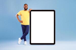 Full length portrait of cheerful young African American guy leaning on giant tablet pc, demonstrating blank screen on blue studio background, free space for online advertisement, mockup for newest app photo