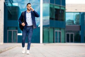 Young middle-eastern guy entrepreneur in formal clothes with backpack going home from office, talking on phone and smiling, having business conversation, business center background, copy space photo