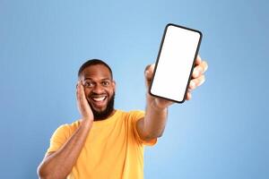 Check out this cool mobile app. Excited African American man presenting smartphone with empty screen, recommending website or ad on blue studio background, space for mockup. Selective focus photo