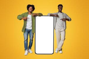 Mobile Ad. Two Black Guys Pointing At Big Smartphone With Blank White Screen, Excited African American Friends Showing Free Copy Space For App Design, Standing Over Beige Studio Background, Mockup photo