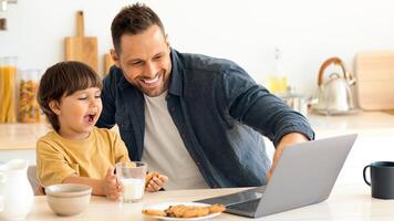 Happy young father with his little son sitting at kitchen and networking on laptop during breakfast, dad showing computer screen to interested kid, free space photo