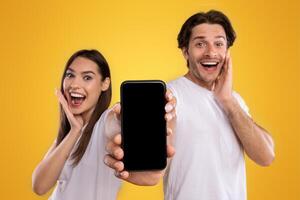 Advertising New Mobile App. Portrait of happy excited couple holding smartphone with black empty screen in hands, showing device with blank space for mock up, saying wow, orange studio background photo