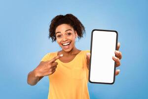Best mobile offer, amazing online sale. Smiling African American lady demonstrating smartphone with mockup, pointing at empty screen on blue background, copy space for app or website design photo