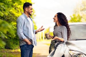 Happy couple having break while travelling by car together, beautiful young middle-eastern man and woman standing by auto at country side, drinking coffee and having conversation, side view photo