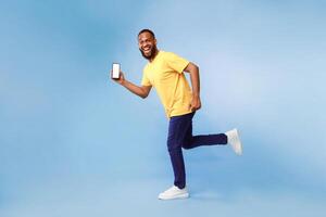Full length of funky young African American guy with blank smartphone running on blue studio background, demonstrating mockup for your mobile app, website or product advertisement photo