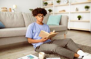 Focused black teenager reading book, sitting on floor, studying from home. Intelligent African American adolescent learning remotely, making assignment, using textbook to get ready for exam or test photo