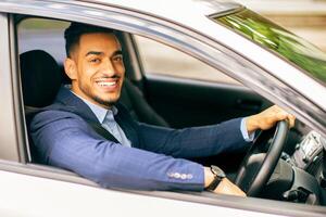 Photo of wealthy middle-eastern businessman in stylish suit driving nice car, going to office, having important business meeting, looking through window and smiling at camera, side view.