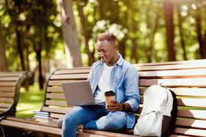 Remote education concept. Cool black student with backpack, books and laptop computer taking part in online class at park. Millennial African American guy participating in web conference outside photo