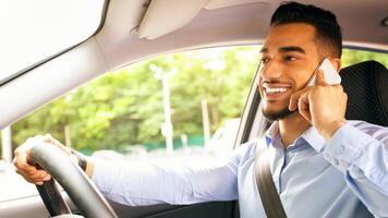 Positive arab guy in formal outfit having phone conversation with lover or business partner while driving car, looking at road and smiling, closeup portrait, side view, copy space. Unsafety driving photo