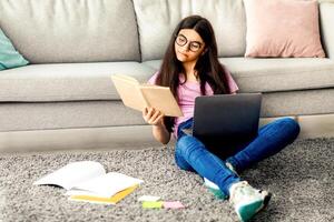 Full length of Indian teen girl reading textbook, sitting on floor with laptop and study materials, learning remotely from home, copy space. Focused adolescent getting ready for online webinar photo