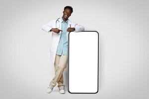 Black Young Male Doctor In Uniform Leaning And Pointing At Big Smartphone With Blank Screen, Showing Free Copy Space For Coronavirus Info Or Vaccination Campaign Design, Standing On Beige Background photo