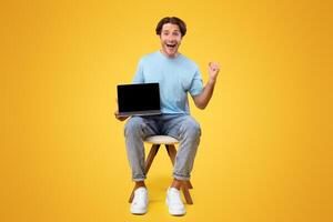 Online Win. Vertical portrait of excited happy guy holding pc laptop with black blank screen, shaking clenched fist making winner gesture, screaming yes showing device with empty free space for mockup photo