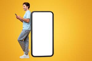 Great App. Excited Man Leaning On Big Smartphone With Blank White Screen And Using His Cell Phone, Cheerful Guy Chatting On Social Media, Standing On Yellow Background, Mock Up Image, Full Body Length photo