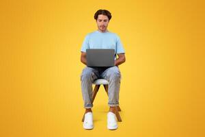 Portrait of serious bearded guy sitting on chair using laptop computer, reading news, watching or movie, working or elearning remotely, isolated on orange studio background, front vertical shot photo