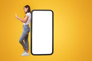 Great App. Happy Lady Leaning On Big Smartphone With Blank White Screen And Using Cell Phone, Cheerful Female Chatting On Social Media, Standing On Orange Background, Mock Up Image, Full Body Length photo