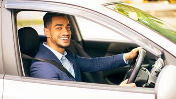 Happy middle-eastern young businessman driving new auto, going to office, shot from outside, side view. Handsome successful arab guy manager going home after good day, looking at camera and smiling photo