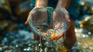 Hands emerge from clear water, holding a fine net filled with lively fish and pebbles. Sunlight dances on the surface, illuminating the vibrant colors of the catch. photo