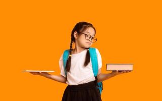 Confused Japanese Schoolgirl Comparing Tablet Computer And Book Standing Over Yellow Background. Books Vs Gadgets. Studio Shot photo
