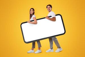Full body length side view profile of cheerful couple carrying big smart phone with blank white screen, walking with gadget under arm, looking at camera isolated on yellow studio background, mock up photo