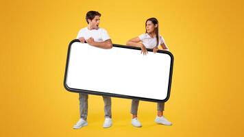 It's Mine. Couple holding big smartphone with white empty screen on yellow orange studio background fighting for gadget pulling device in different directions. Mock up display template, banner photo