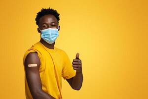 Coronavirus Vaccination Advertisement. Happy Vaccinated Black Guy With Plaster On Arm Showing Thumb Up After Getting Covid-19 Vaccine Injection, African Man In Medical Mask Posing On Beige Background photo