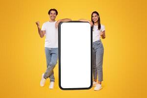 Yes. Excited Couple Leaning On Big Smartphone With Empty White Screen Shaking Clenched Fists, Cheerful Guy And Lady Celebrating Win, Standing On Yellow Background, Mock Up Collage, Full Body Length photo
