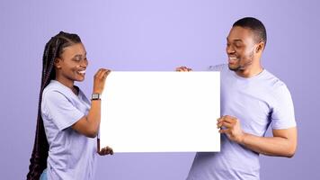 Portrait of cheerful smiling millennial black couple holding and looking at white paper placard for advertisement or text, free copy space for advertising, isolated on violet purple studio background photo